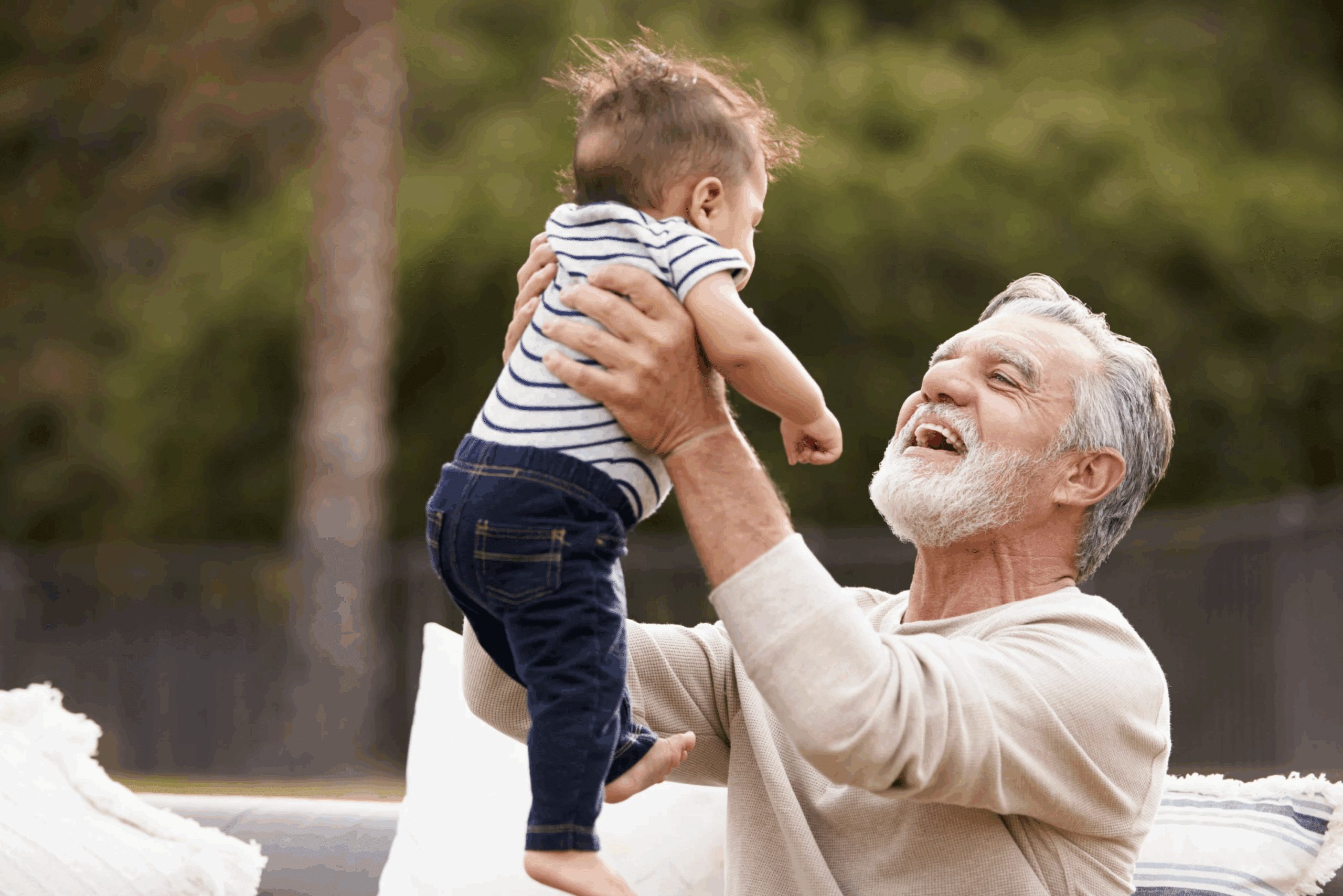 An older man with gray hair and a beard lifts a smiling baby wearing a striped shirt and jeans. They are outdoors on a couch with greenery in the background.