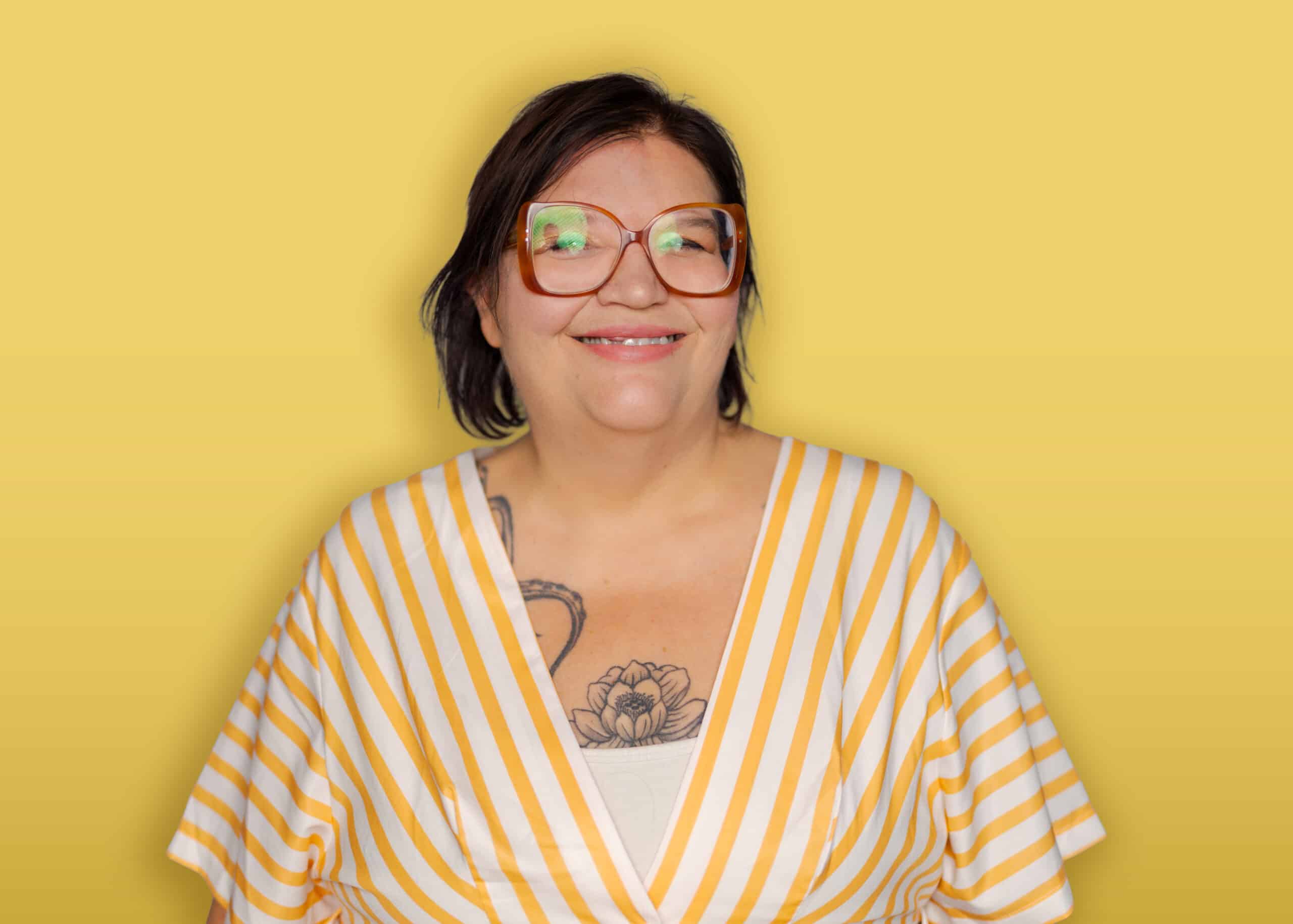 A person with dark hair, wearing a black headband, a green button-up shirt over a black top, and earrings stands in front of a yellow background and smiles at the camera.