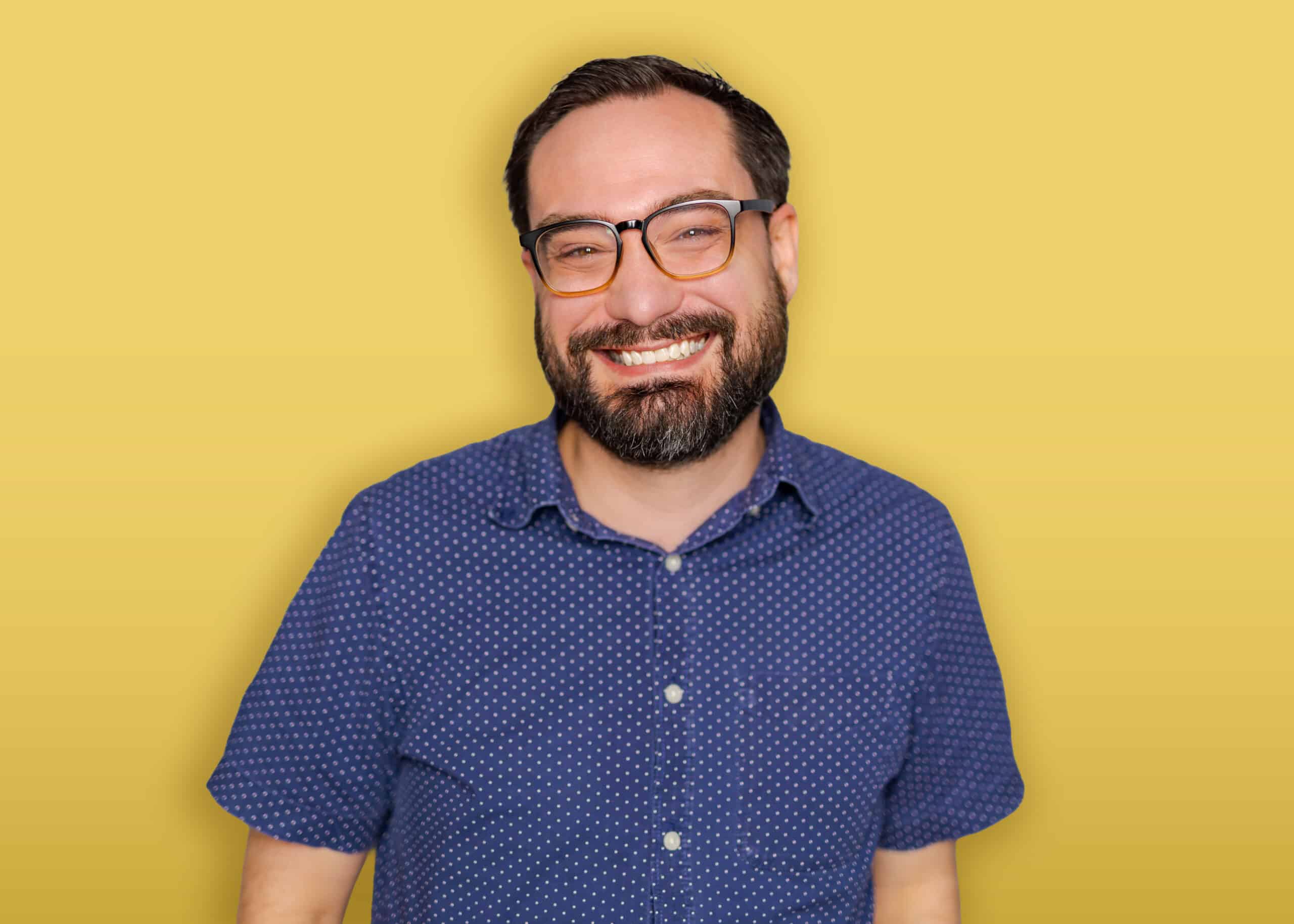A person with dark hair, wearing a black headband, a green button-up shirt over a black top, and earrings stands in front of a yellow background and smiles at the camera.