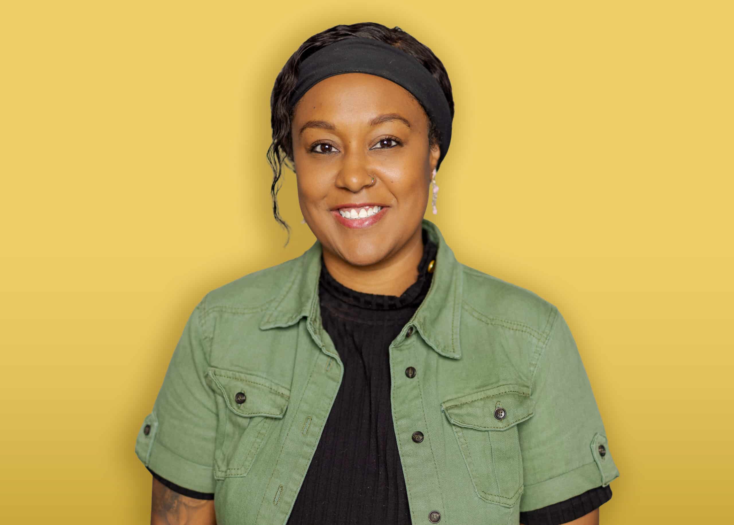 A person with dark hair, wearing a black headband, a green button-up shirt over a black top, and earrings stands in front of a yellow background and smiles at the camera.