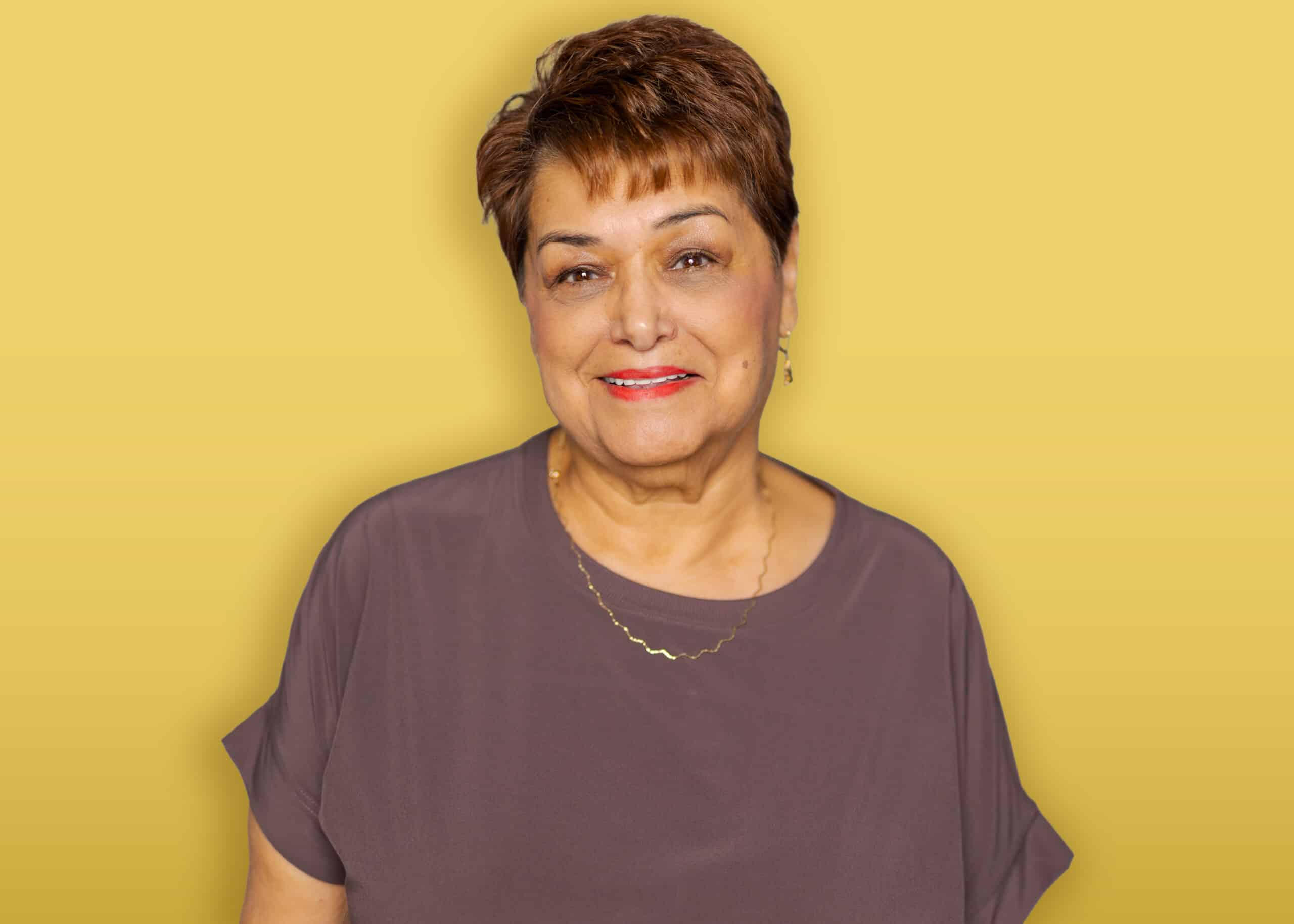 A person with dark hair, wearing a black headband, a green button-up shirt over a black top, and earrings stands in front of a yellow background and smiles at the camera.