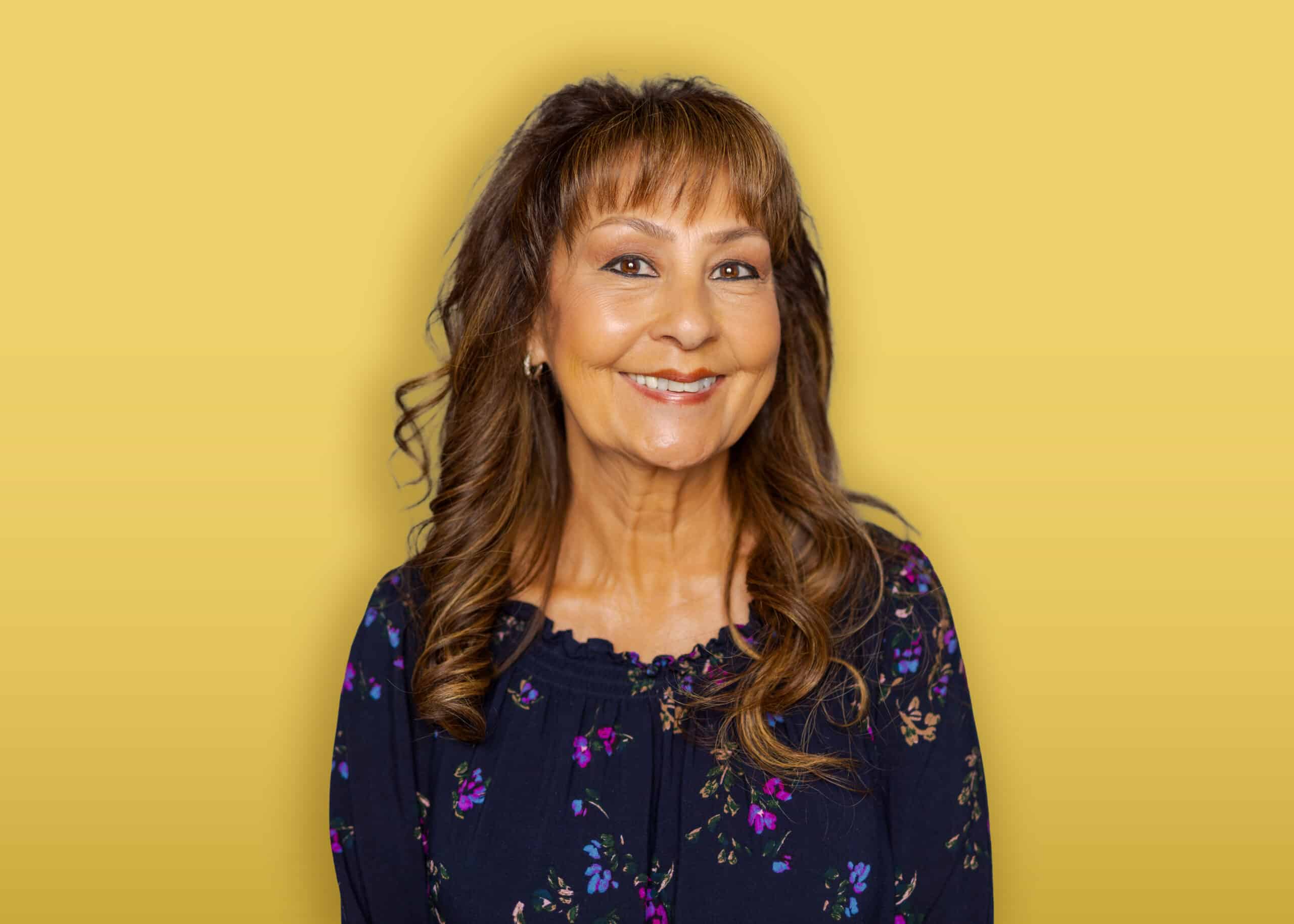 A person with dark hair, wearing a black headband, a green button-up shirt over a black top, and earrings stands in front of a yellow background and smiles at the camera.