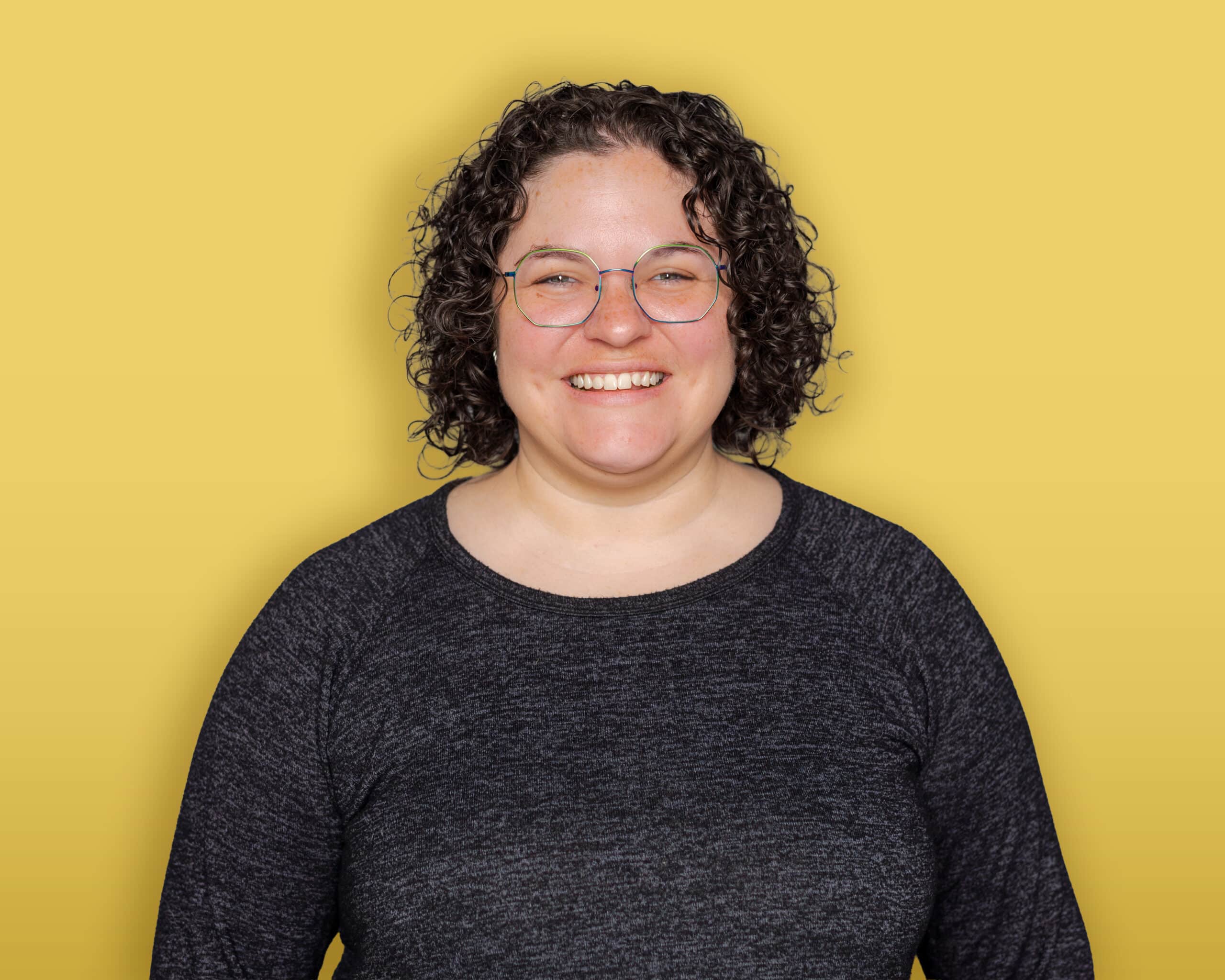 A person with dark hair, wearing a black headband, a green button-up shirt over a black top, and earrings stands in front of a yellow background and smiles at the camera.