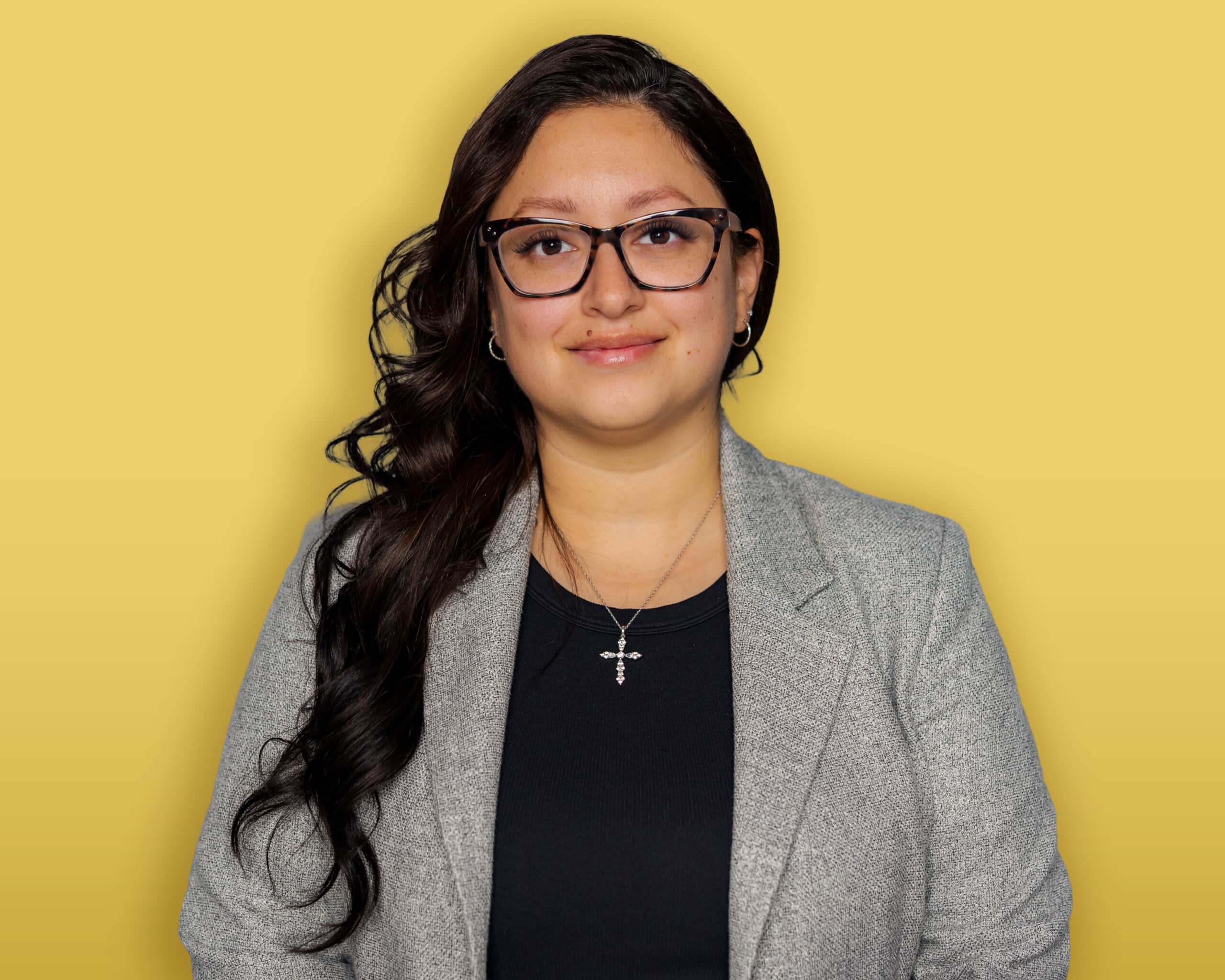 A person with dark hair, wearing a black headband, a green button-up shirt over a black top, and earrings stands in front of a yellow background and smiles at the camera.