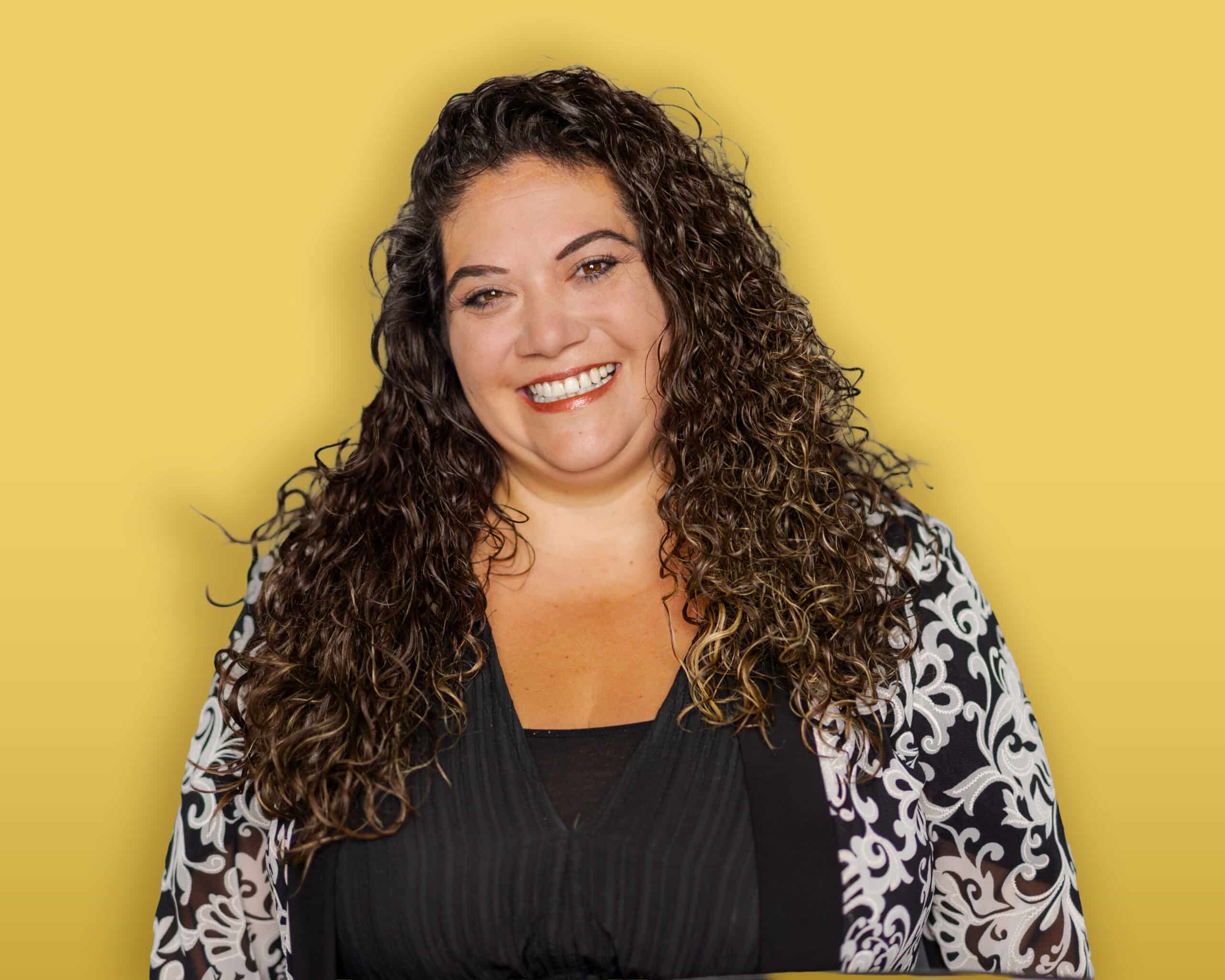 A person with dark hair, wearing a black headband, a green button-up shirt over a black top, and earrings stands in front of a yellow background and smiles at the camera.