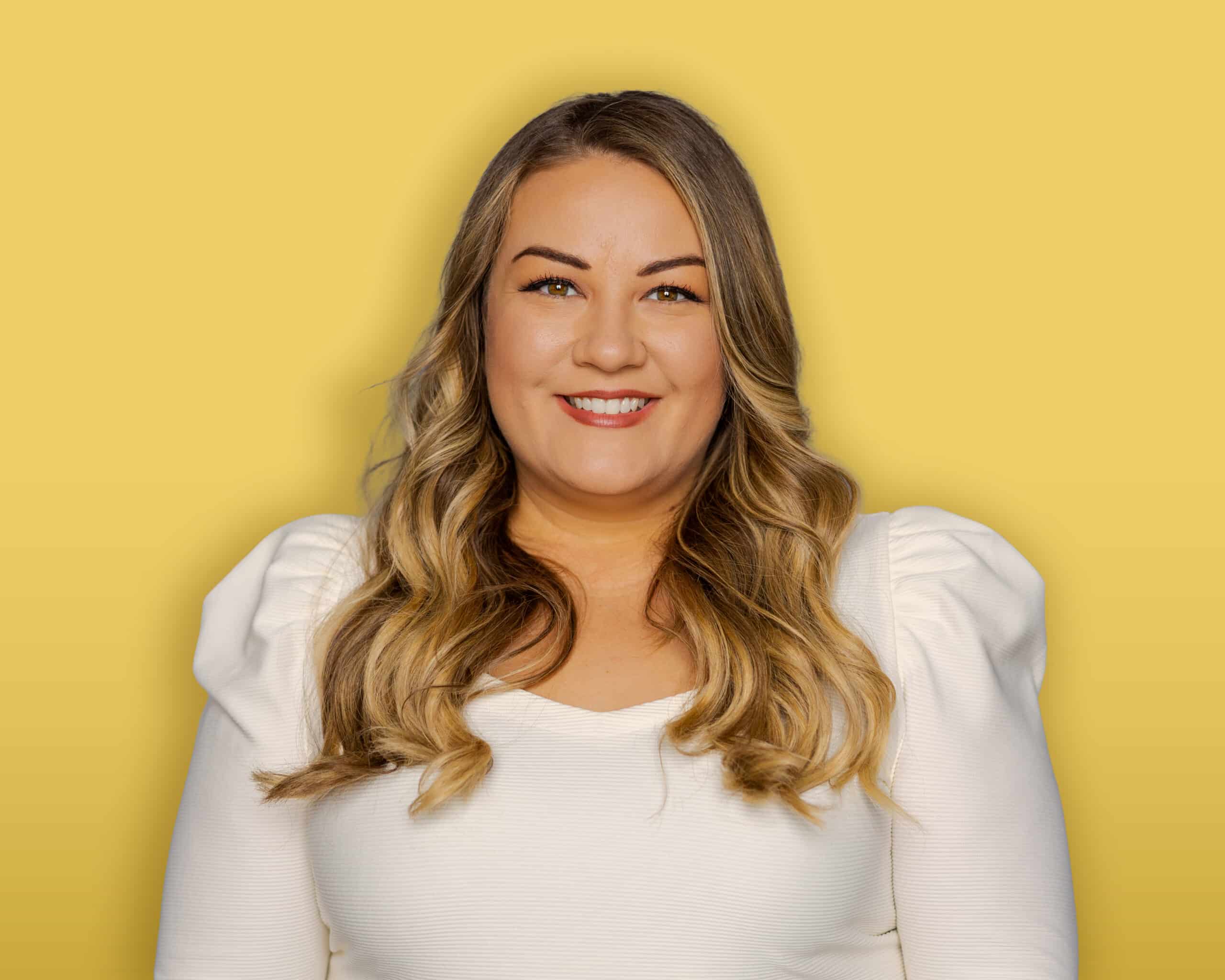 A person with dark hair, wearing a black headband, a green button-up shirt over a black top, and earrings stands in front of a yellow background and smiles at the camera.