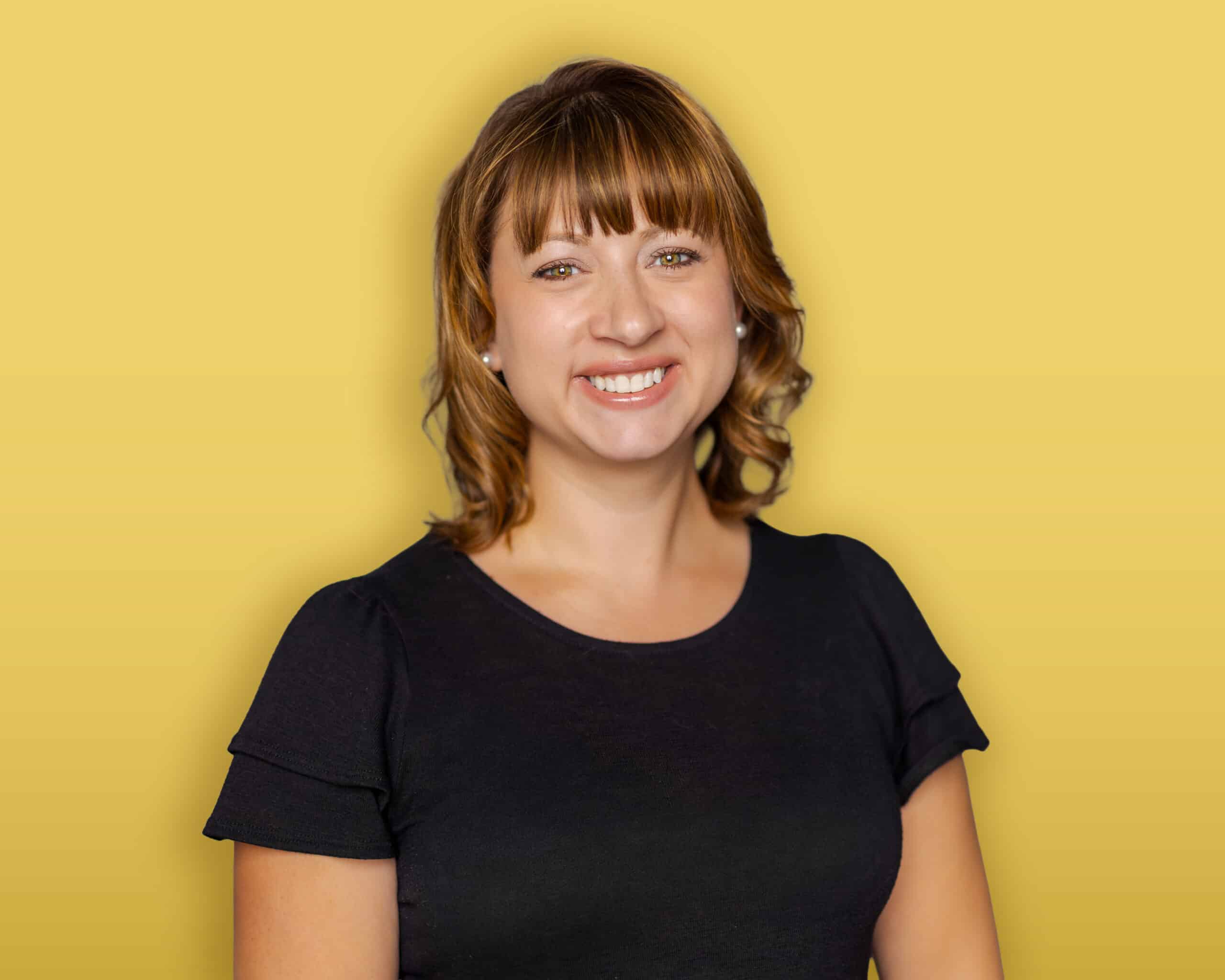 A person with dark hair, wearing a black headband, a green button-up shirt over a black top, and earrings stands in front of a yellow background and smiles at the camera.
