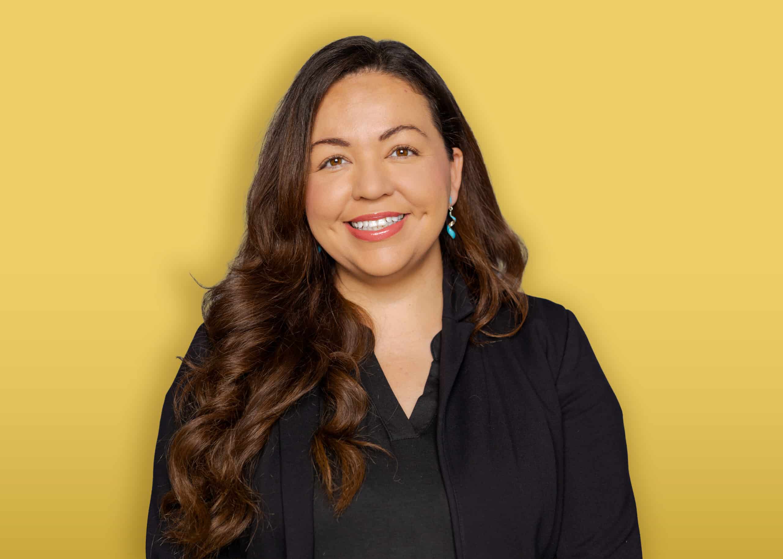 A person with dark hair, wearing a black headband, a green button-up shirt over a black top, and earrings stands in front of a yellow background and smiles at the camera.