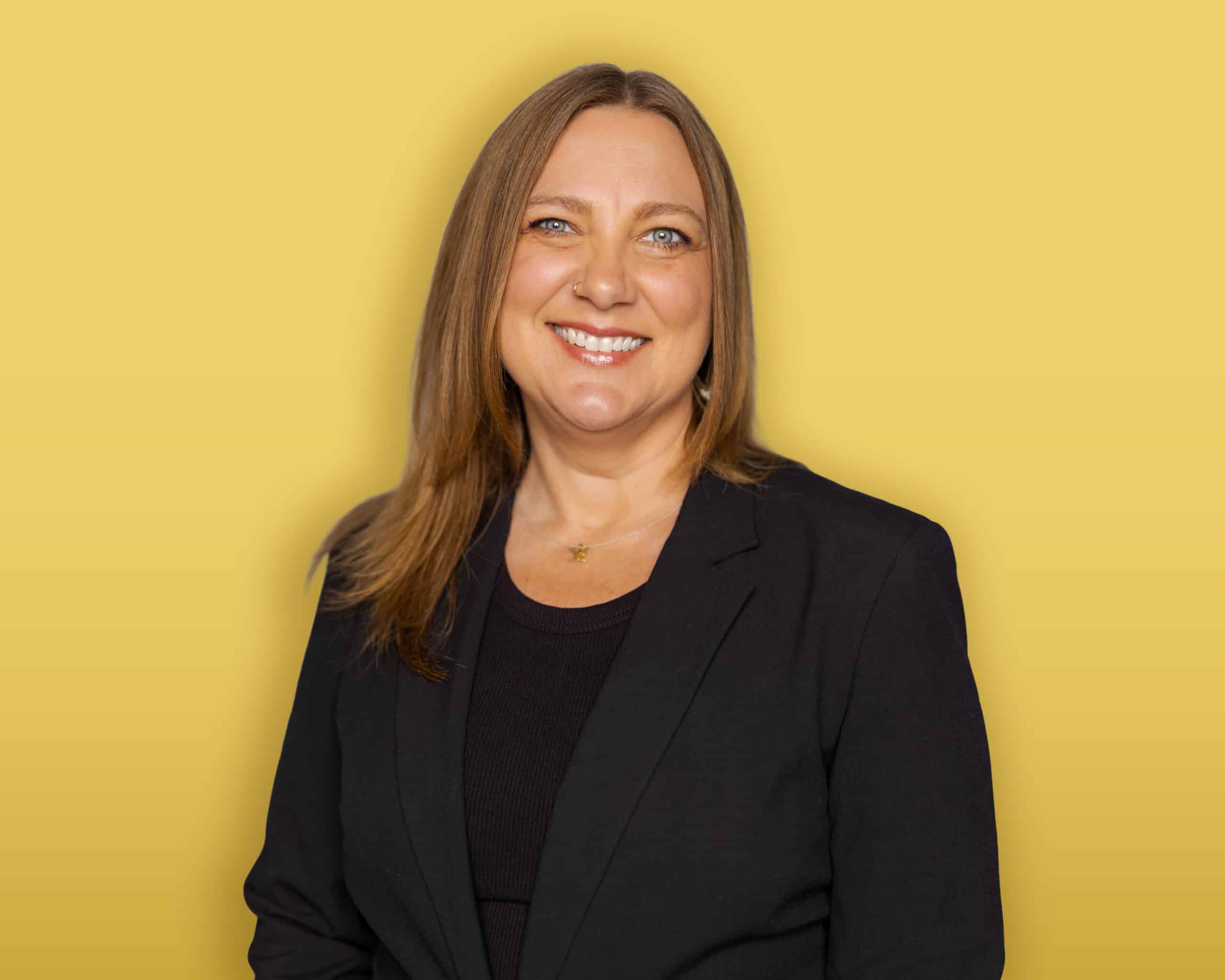 A person with dark hair, wearing a black headband, a green button-up shirt over a black top, and earrings stands in front of a yellow background and smiles at the camera.