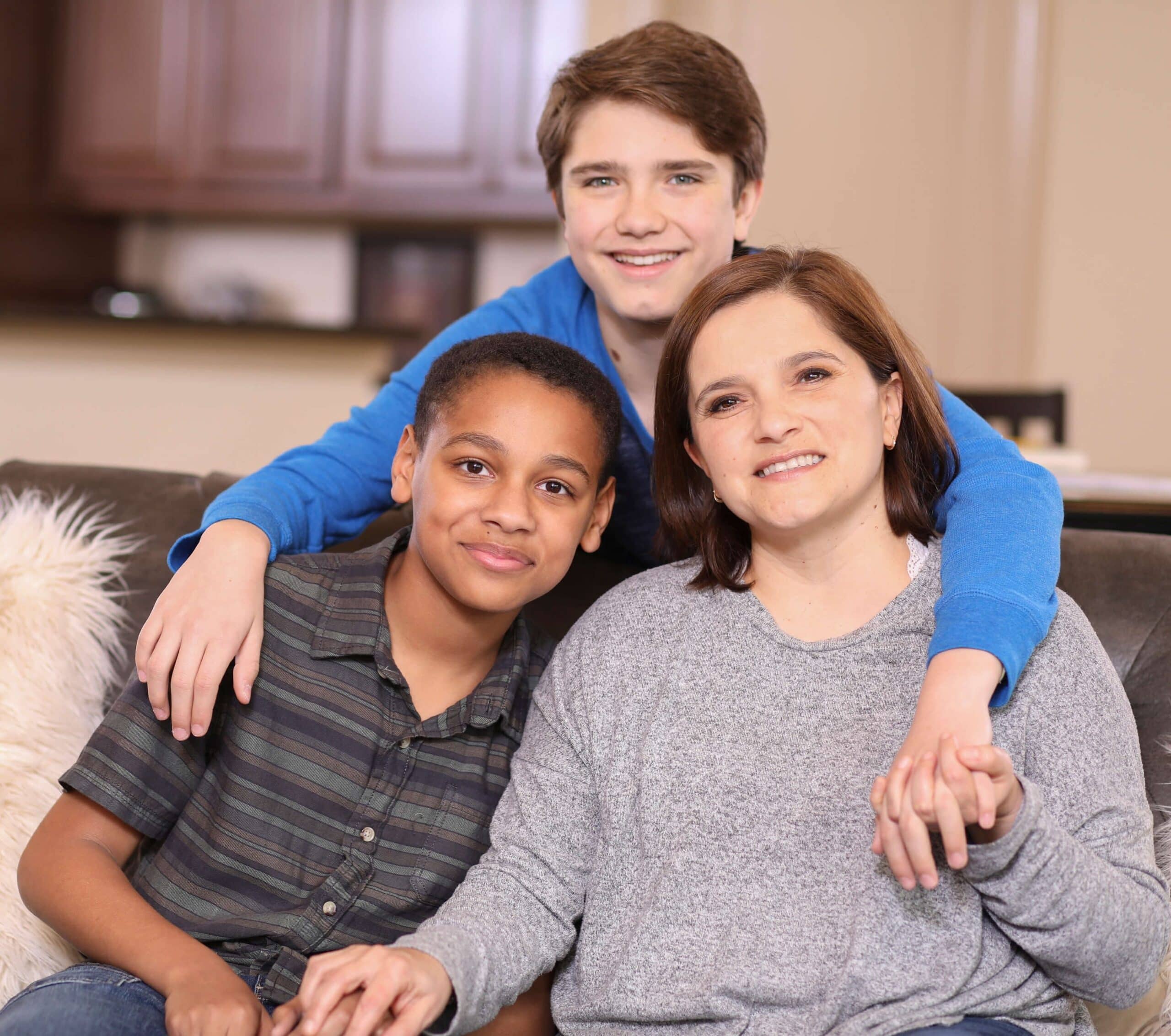 Three people sit close together on a couch, smiling at the camera. An adult holds hands with two children, one sitting beside them and the other standing behind with arms around their shoulders. The setting appears to be a home.