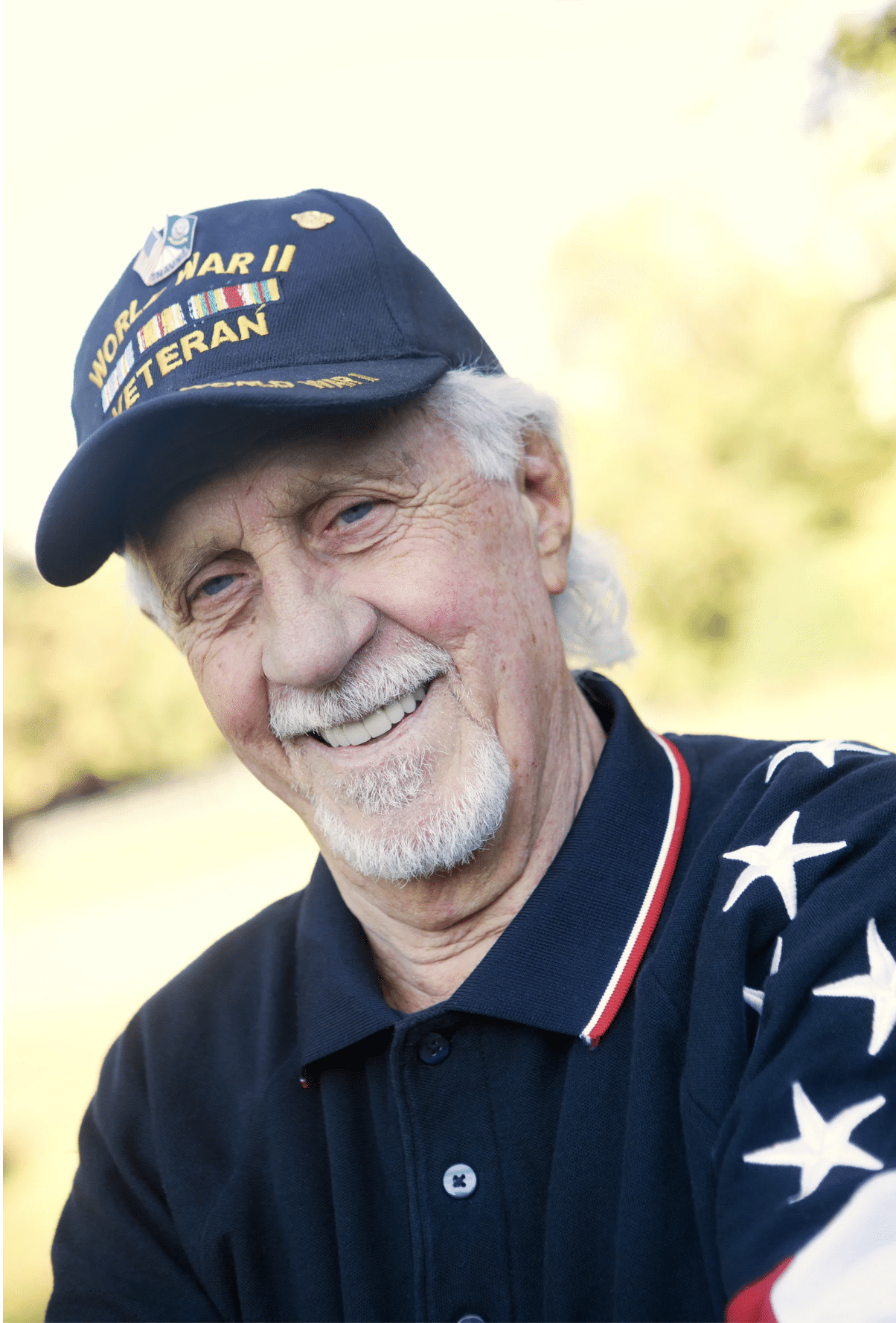 An older man with gray hair and a beard wears a World War II Veteran hat and a navy shirt with white stars on the sleeve. He is outdoors and smiling at the camera.