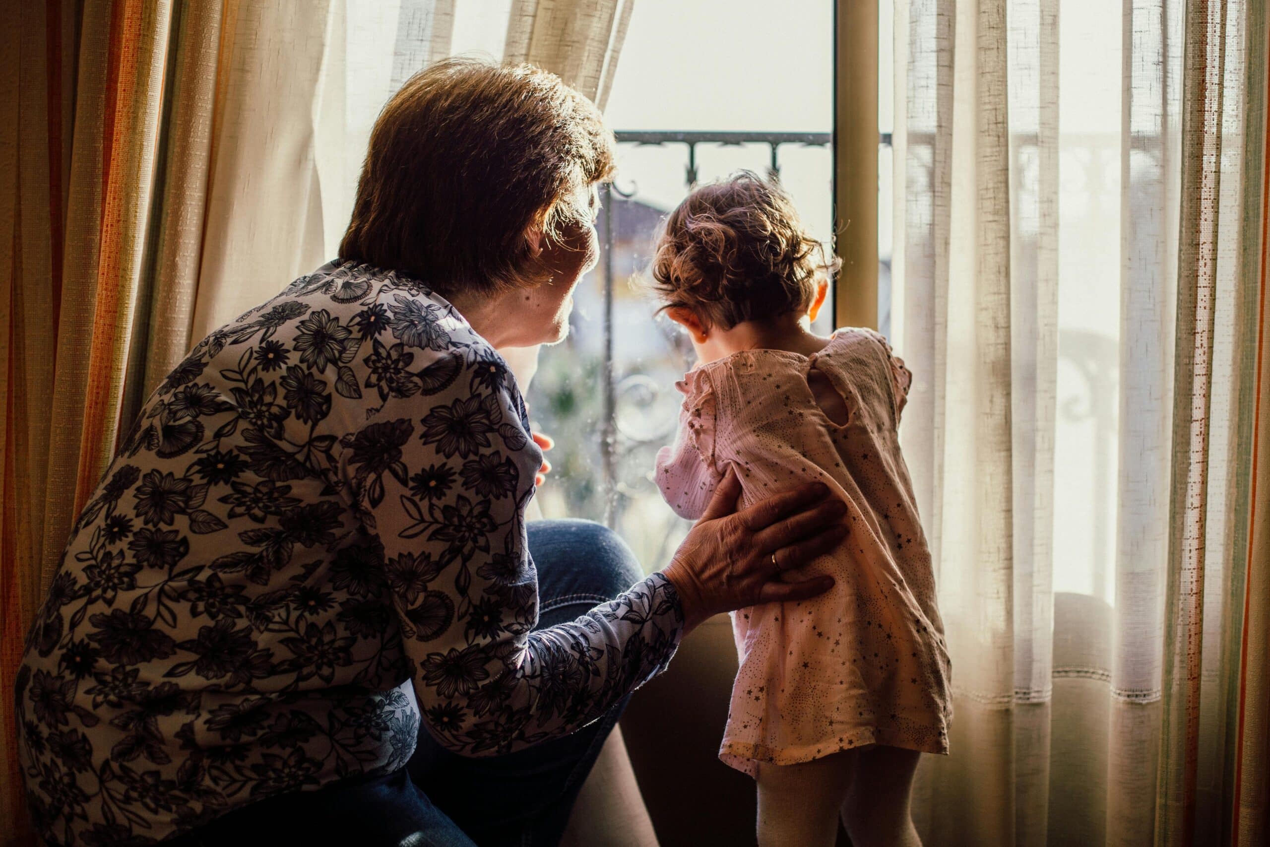 An adult and a young child stand by a window with sheer curtains. The adult has a hand on the child’s back as they both look outside, with sunlight streaming in through the glass.