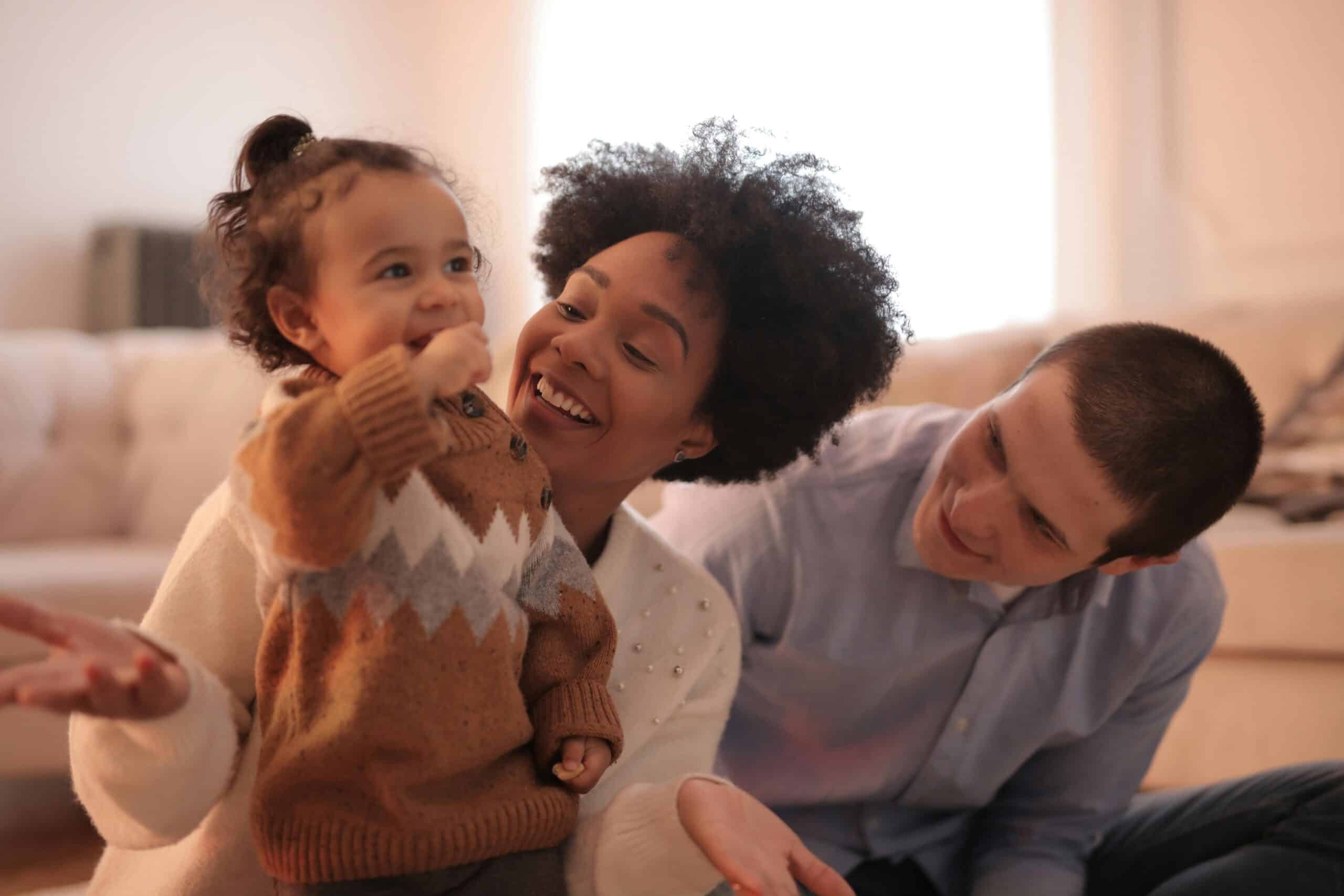 A small child in a brown sweater stands in front of two adults on a couch. The child is smiling with one hand raised. The adults, one woman and one man, are looking at the child and smiling.