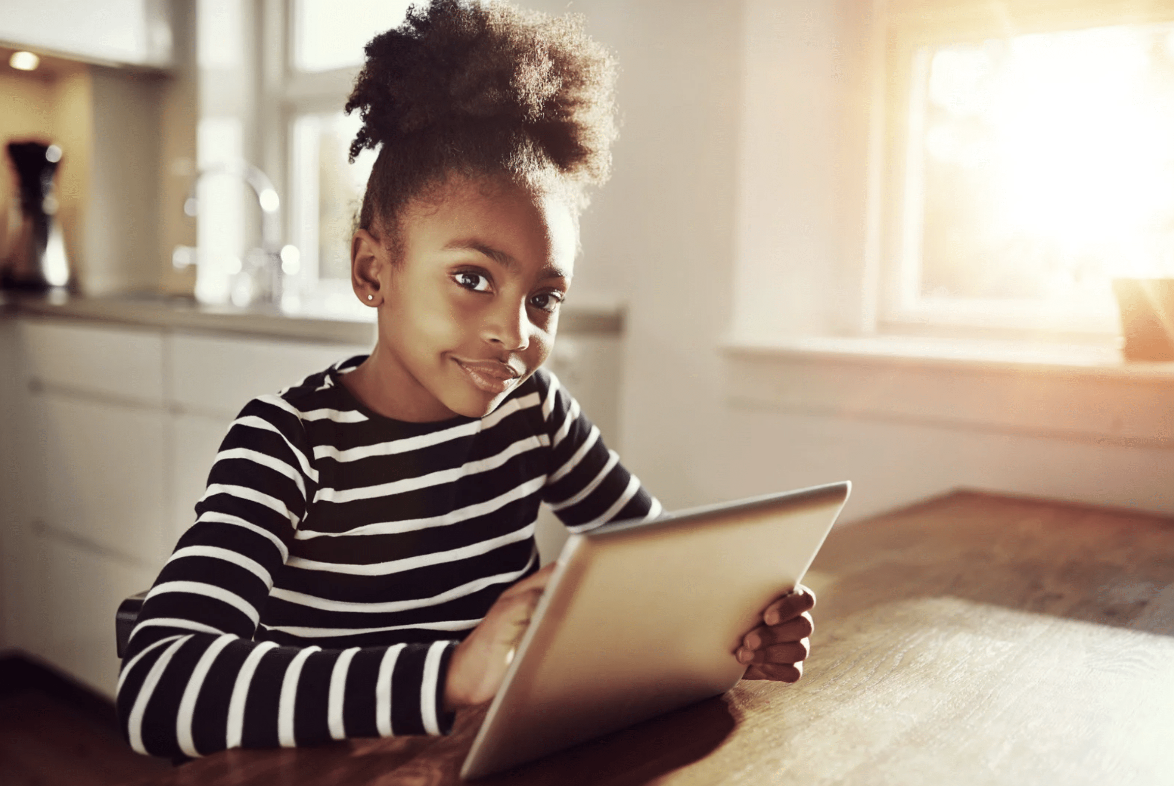 A young girl wearing a black and white striped shirt sits at a wooden table in a sunlit kitchen, holding and using a tablet.