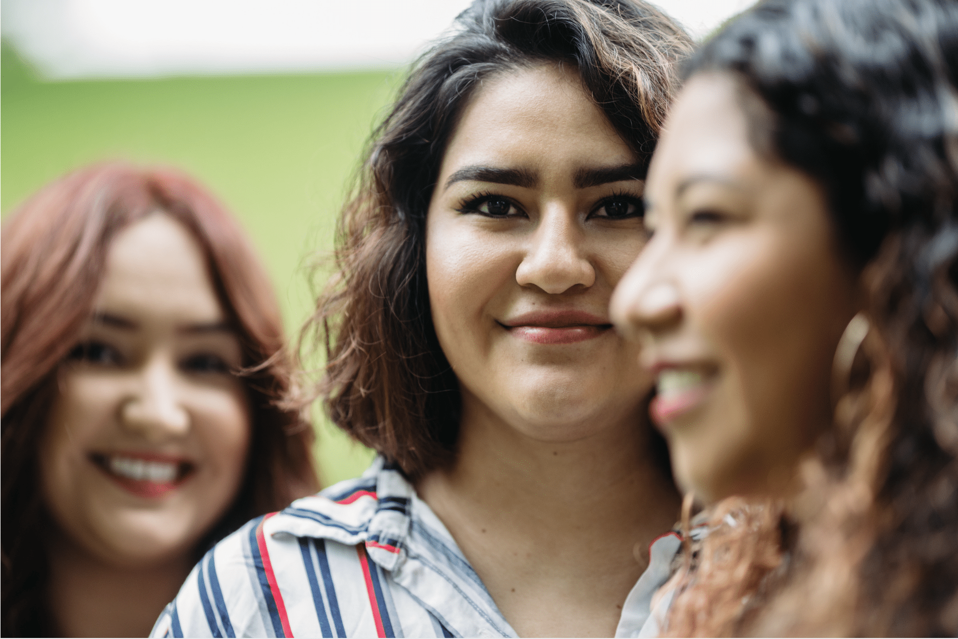 Three women are outdoors, standing close together. The woman in the center is in focus and looking directly at the camera, while the two women on either side are smiling and slightly out of focus. The background is green.