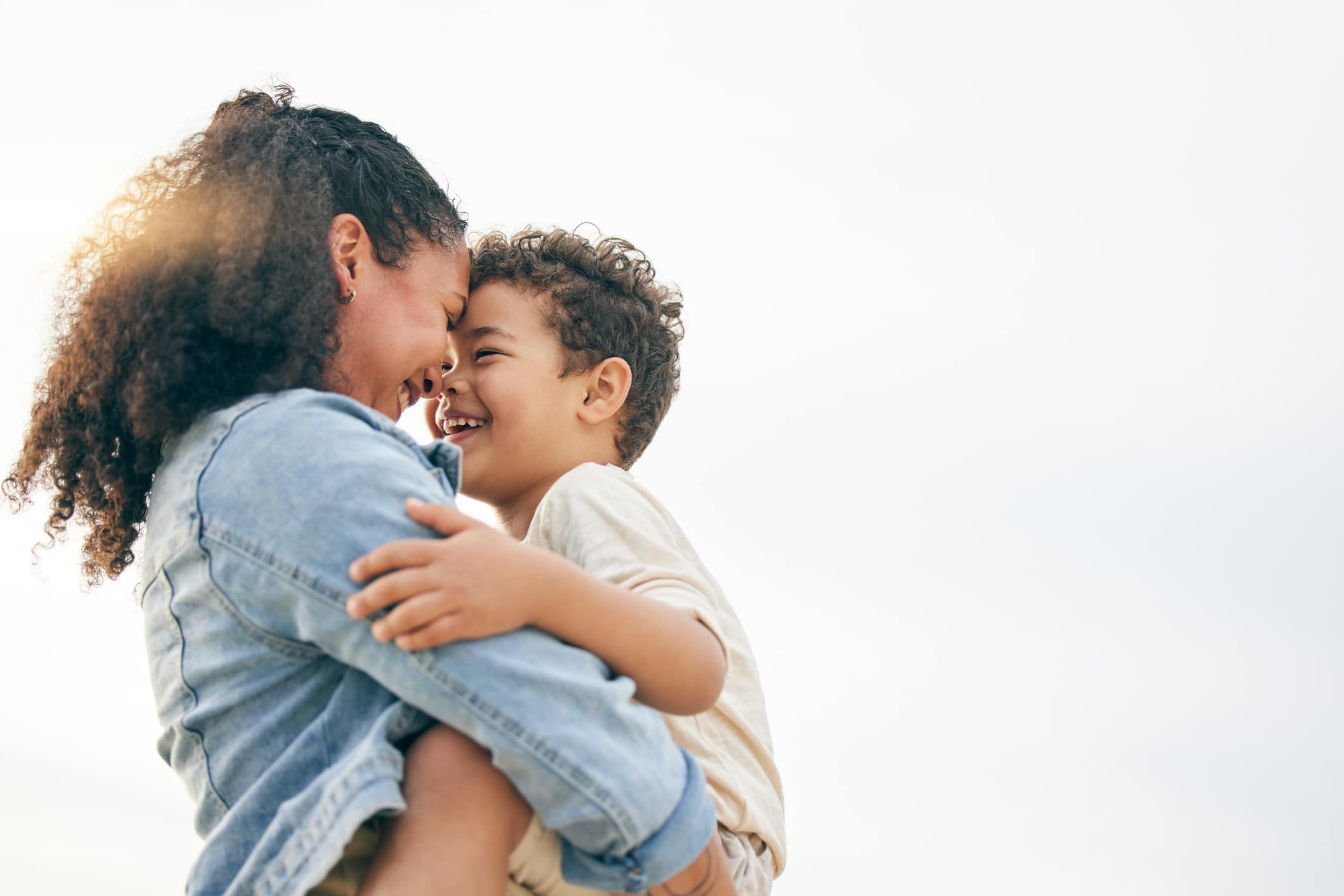 A woman and a young boy face each other, smiling and touching foreheads. The woman is wearing a denim jacket and the boy is wearing a light-colored shirt. The background is bright and clear.
